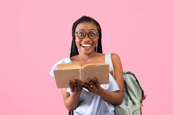 Aufgeregte junge afroamerikanische Studentin mit Rucksack, aufgeschlagenem Buch auf rosa Hintergrund - Foto, Bild