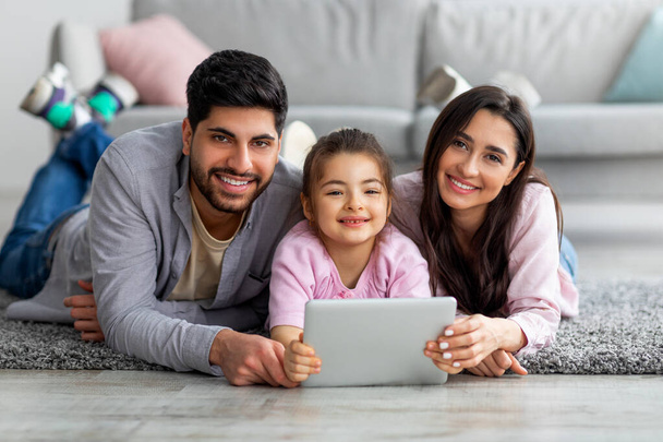 Young arab parents and their cute daughter using digital tablet while lying on floor together and smiling to camera - Photo, Image
