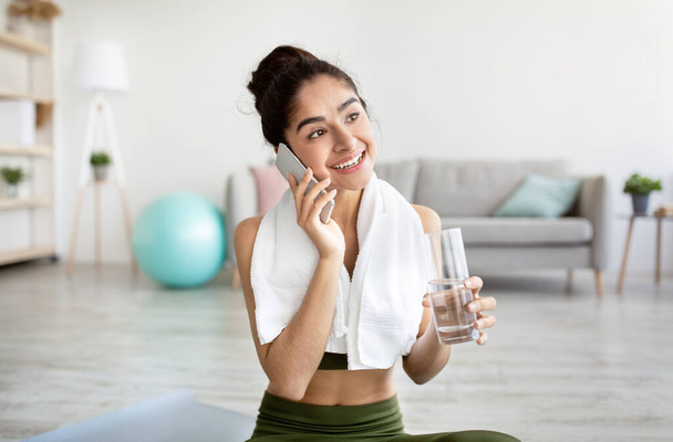 Fit young Indian woman holding glass of water, talking on smartphone, having break from domestic training - 写真・画像