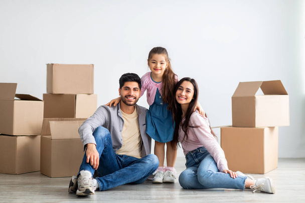 Cheerful eastern family of three people moving to new house, sitting among cardboard boxes in empty living room - Foto, Bild