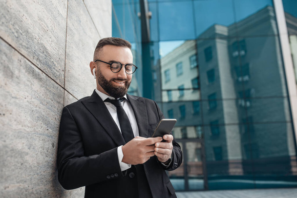 Cesser le travail. Homme d'affaires mature souriant en vêtements formels utilisant un smartphone et écoutant de la musique près de l'immeuble de bureaux - Photo, image