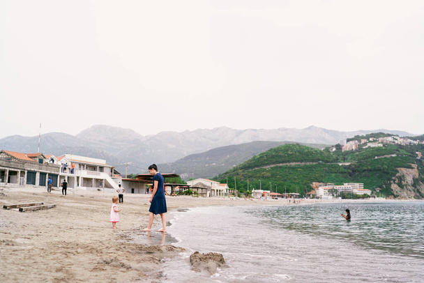 山や緑や建物を背景に海沿いの砂浜に立つ母と少女 - 写真・画像