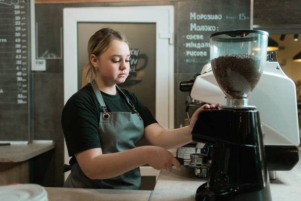 バリスタは、カフェでコーヒーグラインダーで豆を挽くと、ポートフィルターを保持しています。彼の手の中にポータフィルターで悲しい女の子のウェイター。コーヒーマシンのためのコーヒーを準備するプロセス. - 写真・画像