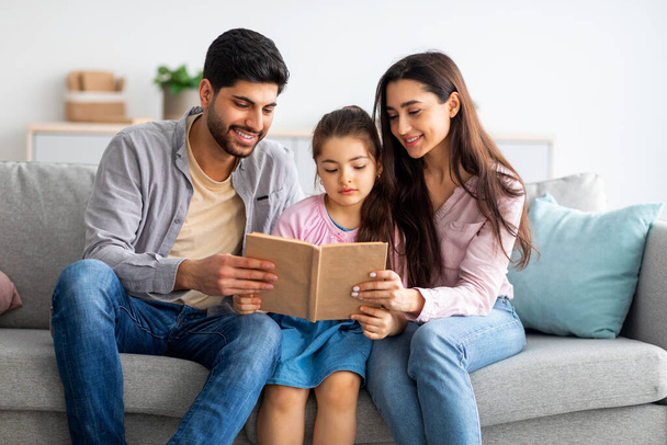 Rodinná zábava. Šťastné arabské rodiče čtení knihy se svou dcerou, zatímco relaxaci společně na pohovce - Fotografie, Obrázek