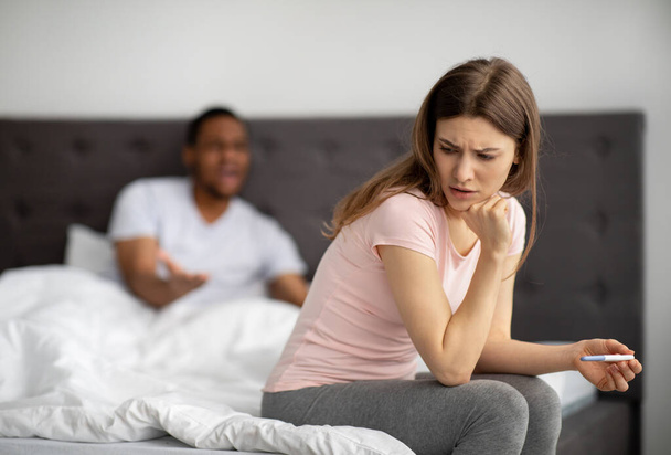 Nieszczęśliwa młoda kobieta siedząca na łóżku z testem ciążowym, czarny mąż krzyczący na nią, wewnątrz - Zdjęcie, obraz