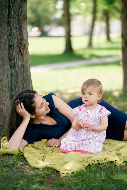 Mom lies next to a little girl sitting on a blanket on the green grass - Foto, Imagem