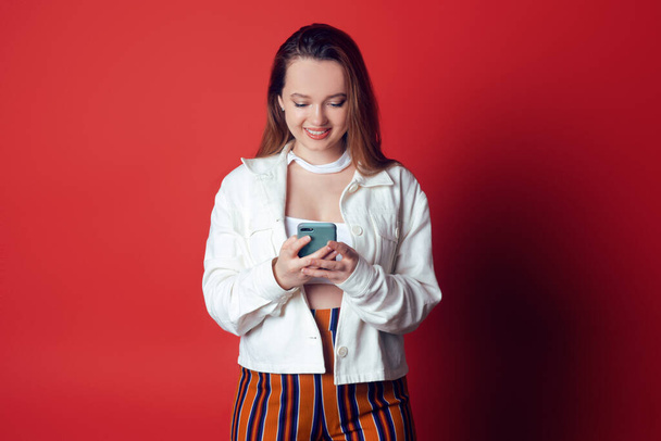 Beautiful, sweet, brunette on a red background. The girl looks into the phone and laughs, smiles. Empty space for text. A woman with beautiful hair and a smile is standing with a mobile phone. - Photo, Image
