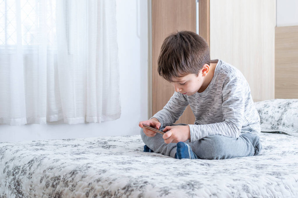 6 yaşındaki küçük bir çocuk akıllı telefon kullanıyor ve oynuyor. Küçük tatlı çocuk yatakta akıllı telefonda oyun oynuyor. Çocuk akıllı telefonuyla video oyununda oynuyor. Sosyal ve teknoloji kavramı. - Fotoğraf, Görsel