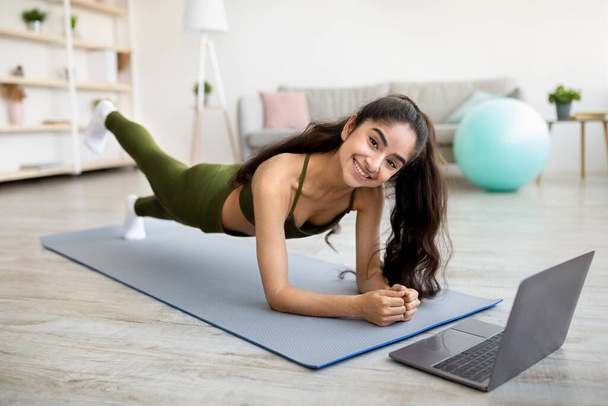 Mujer india fuerte de pie en el tablón del codo delante de la computadora portátil, ejercitando a tutorial de deportes en línea en casa - Foto, Imagen