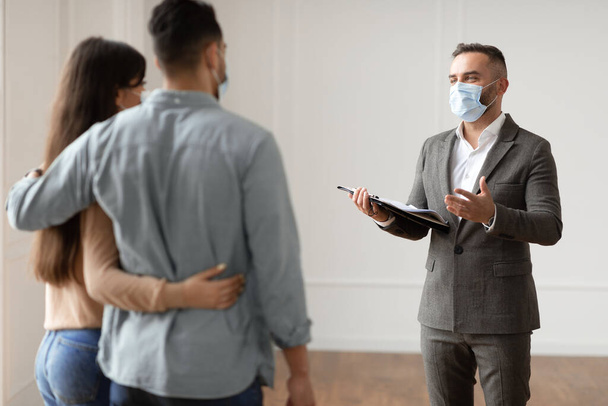 Maschio agente immobiliare in maschera medica che mostra agli acquirenti nuovo appartamento - Foto, immagini