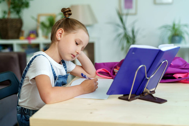 little girl is tired of learning. home schooling, homework. girl rubs his eyes from fatigue reading books and textbooks. little kid student sitting at table with books. vision problems - Photo, Image