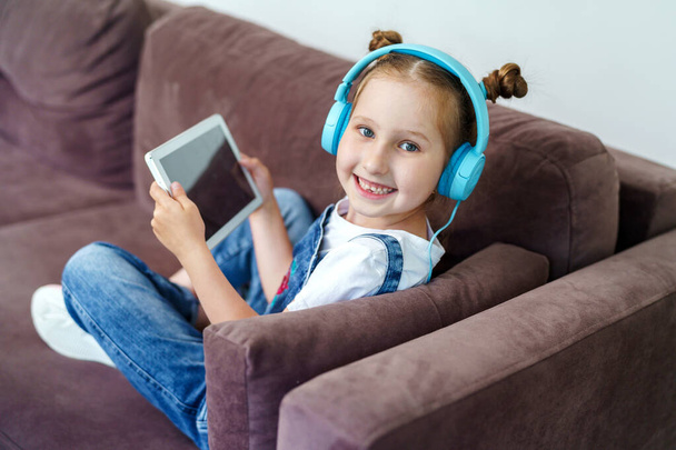 pequeña linda chica con auriculares y una tableta está sentado en casa en el sofá. El niño está feliz de aprender en casa en línea en una tableta de computadora, escuchando música. Una nueva aplicación de aprendizaje para niños. - Foto, imagen