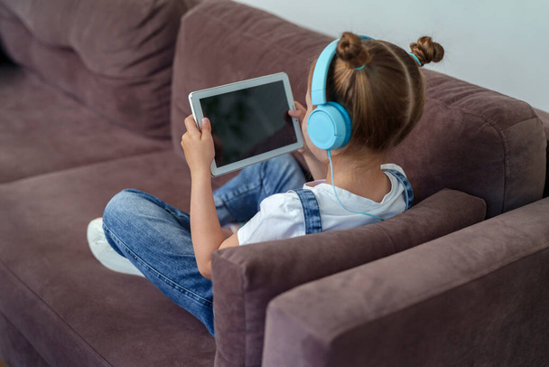 little cute girl with headphones and a tablet is sitting at home on the couch. The child is happy to learn at home online on a computer tablet, listening to music. A new children's learning app. - Foto, Bild