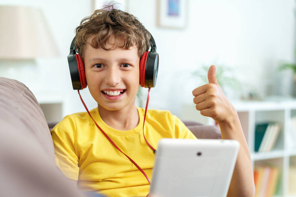 happy boy with headphones and tablet shows thumbs up, sitting at home on the couch. the child is happy to learn at home online on computer tablet, listening to music. a new app for teaching children. - Zdjęcie, obraz