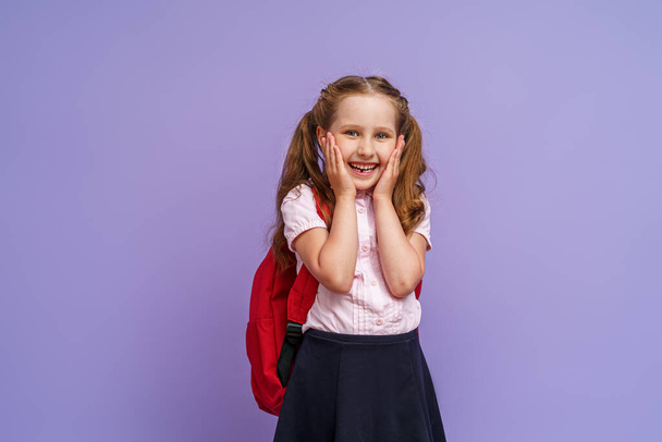 mischievous laughing schoolgirl in a uniform with a backpack, stands on a purple background. Back to school. the child is very excited and happy. A little girl begins to study, to learn. Advertising. - Φωτογραφία, εικόνα