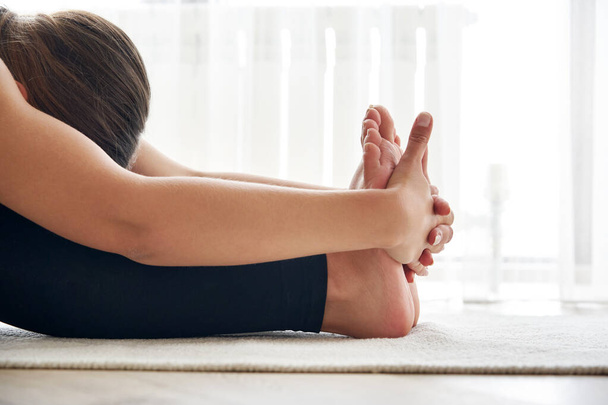 Обрезанный вид на молодую женщину практикующую йогу, сидящую в сидящих вперед изгиб упражнения, paschimottanasana позе - Фото, изображение