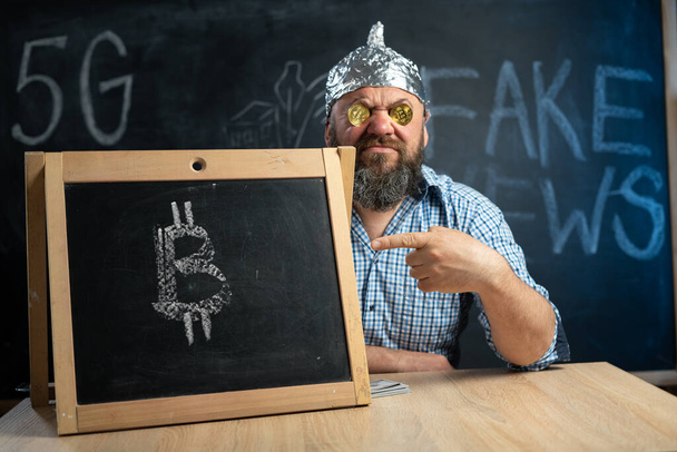 bitcoin cryptocurrency coins in the eyes of a conspiracy theorist sitting at the table holds a chalk board with a bitcoin sign in his hands. Crazy man in a shirt with crypto currency money. - Photo, Image