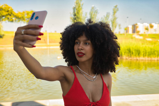 Bella giovane donna afro americana con capelli afro con top in corallo e gonna bianca che si fa un selfie. - Foto, immagini