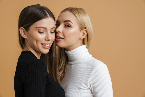 Two attractive smiling young sensual women embracing while standing isolated over beige background - Foto, Bild