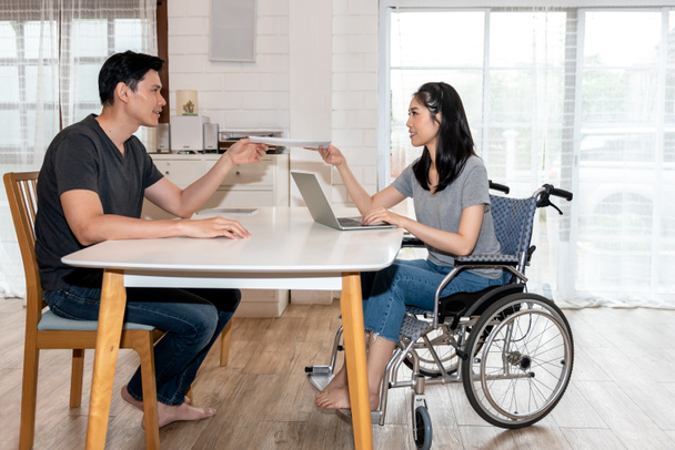 Ασιατικό νεαρό ζευγάρι, οι γυναίκες είναι ασθενείς που κάθονται σε αναπηρική καρέκλα, εργάζονται μαζί χρησιμοποιώντας ένα φορητό υπολογιστή, έννοια για την υγειονομική περίθαλψη και οι άνθρωποι των επιχειρήσεων εργάζονται από το σπίτι - Φωτογραφία, εικόνα