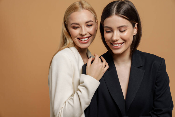 Two beautiful smiling elegant women wearing smart jackets posing together isolated over beige background - Fotoğraf, Görsel