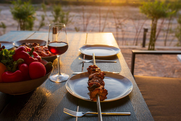 Barbecue on the terrace. A picnic on the terrace for friends. Terrace with grill, meat at sunset. Fried meat in the open air at home. Cooking at home in the summer on the street. Kebab. Grilled meat at home set the table.Meat with wine and vegetables - Photo, Image
