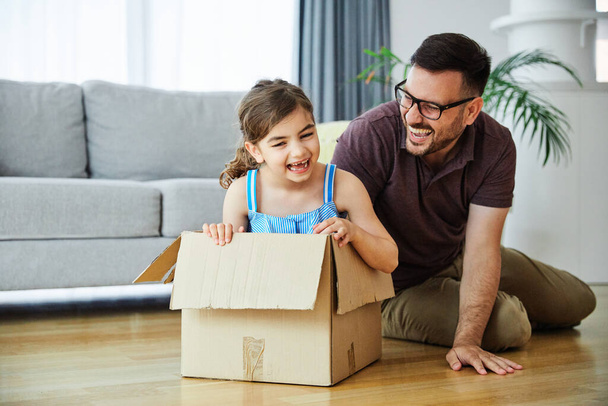 Ojciec popycha córkę do siedzenia w kartonowym pudełku, bawiąc się w domu - Zdjęcie, obraz