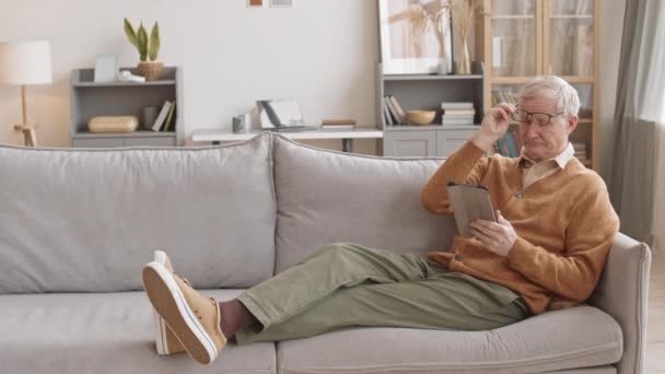 Slowmo des älteren Mannes in Freizeitkleidung und Brille mit digitalem Tablet beim Entspannen auf dem Sofa in der hellen, gemütlichen Wohnung - Filmmaterial, Video