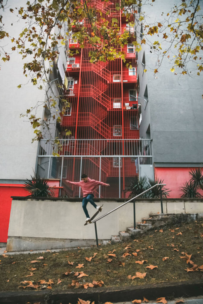 Łyżwiarz w różowej bluzie na desce węży szlifujący poręcz miejskiej ulicy z czerwono-szarym budynkiem w tle. Deskorolkarz w akcji. Miejska koncepcja. Przestrzeń kopiowania - Zdjęcie, obraz
