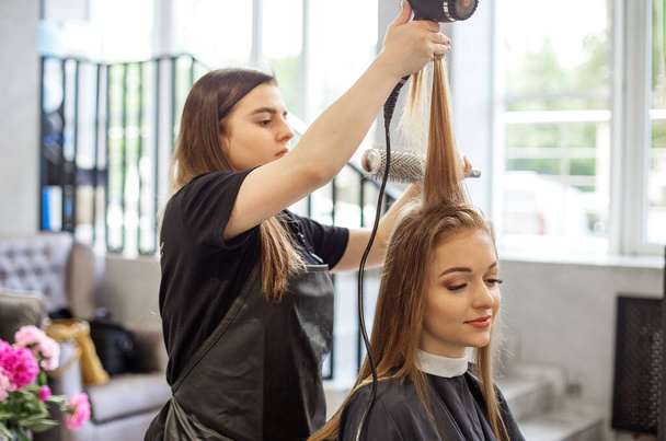 Uma menina em um salão de beleza, um cabeleireiro faz seu cabelo, corta seu cabelo para uma menina com cabelo comprido. uma mulher pinta o cabelo, seca o cabelo com um secador de cabelo. - Foto, Imagem