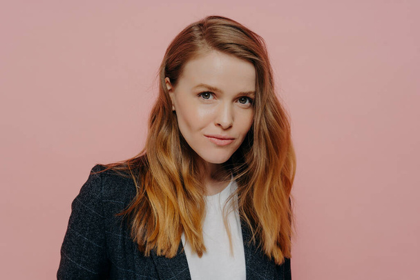 Attractive young woman with ginger hair in formal jacket posing against pink background - Photo, image