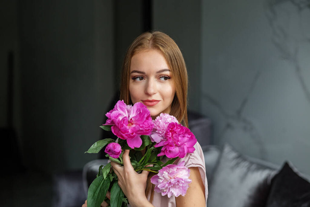 A beautiful girl with long blonde hair in a modern interior holds a bouquet of pink peonies in her hands, a romantic girl. - Photo, Image