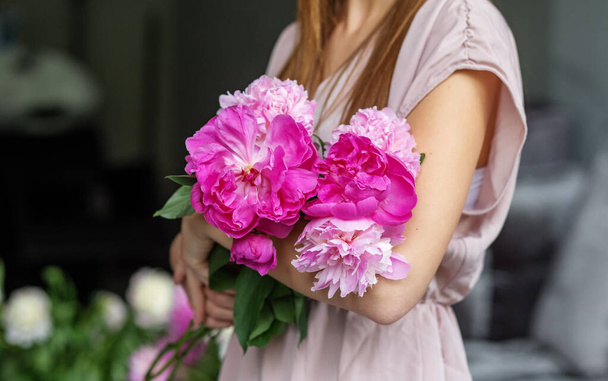 Een mooi meisje met lang blond haar in een modern interieur houdt een boeket roze pioenen in haar handen, een romantisch meisje. - Foto, afbeelding
