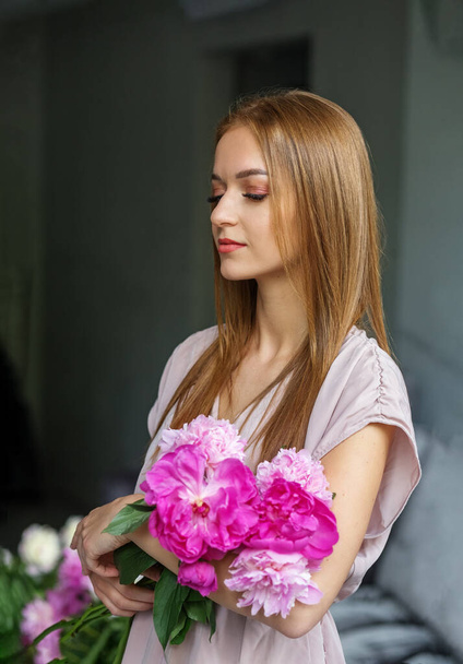 Ein schönes Mädchen mit langen blonden Haaren in einem modernen Interieur hält einen Strauß rosa Pfingstrosen in ihren Händen, ein romantisches Mädchen. - Foto, Bild