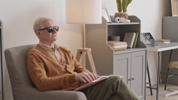 Medium slowmo of blind senior man in casualwear and black eyeglasses reading Braille book while sitting in chair at home - Materiał filmowy, wideo