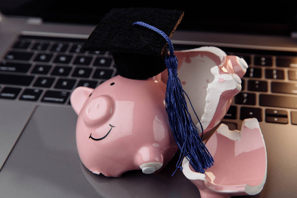Broken pink piggy bank in cap on laptop. College, graduate, diploma concept. Savings for education - 写真・画像