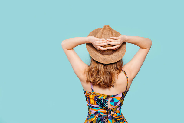 Unknown blonde girl with her back turned wearing a straw hat and a very colourful dress on a blue background. Summer concept. Copy space - Photo, Image