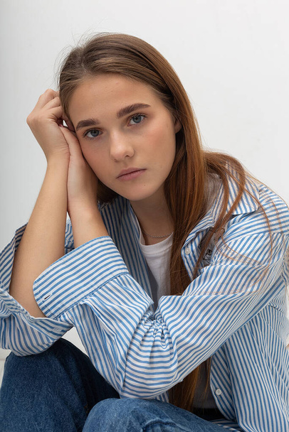 jong Kaukasisch mooi meisje met lang haar in blauw gestreept shirt zit in studio - Foto, afbeelding