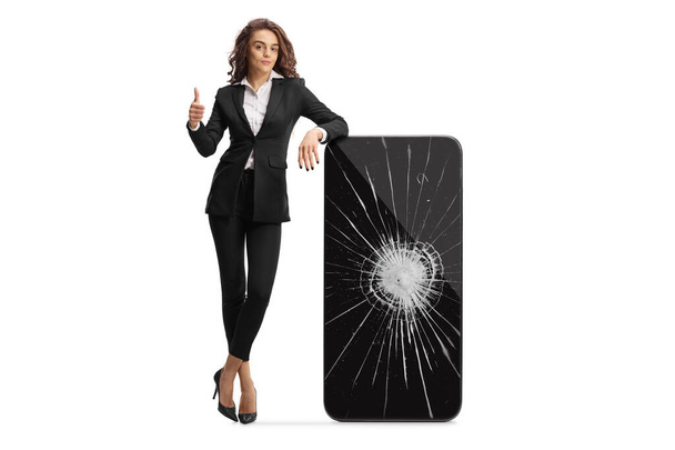 Businesswoman showing thumbs up and leaning on a smartphone with a cracked screen isolated on white background - Photo, Image