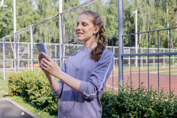 A girl in a sports uniform on the workout site in headphones prints a message on the phone - Фото, изображение