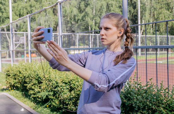 A girl in a sports uniform on the workout site in headphones and a phone in her hands is talking by video call with a pensive expression on her face - Foto, Bild
