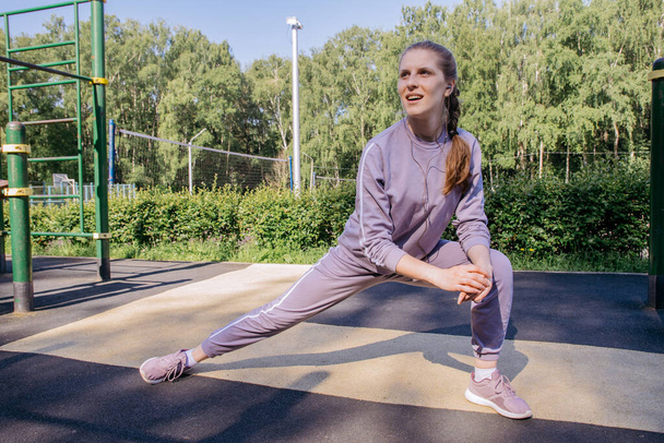 A girl in headphones and a sports uniform on the playground performs a leg stretch - Zdjęcie, obraz