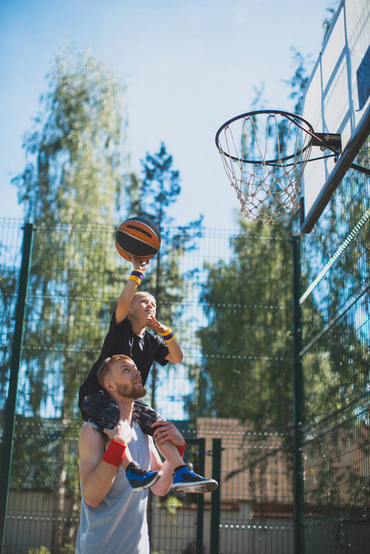 Happy family, young sportive father holding son on shoulders. Kid boy throwing ball in basketball hoop outdoors - Φωτογραφία, εικόνα