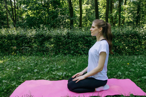 A girl in a white T-shirt and black leggings sits on a pink mat on a summer green lawn - Photo, Image