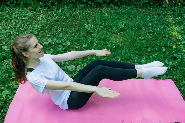 A girl in a white T-shirt and black leggings on a pink mat on a summer green lawn and shakes the press - Foto, imagen