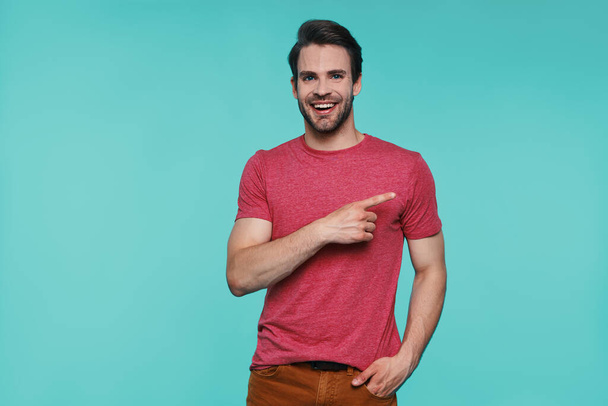 Handsome young smiling man in casual clothing looking at camera and pointing away - Foto, afbeelding