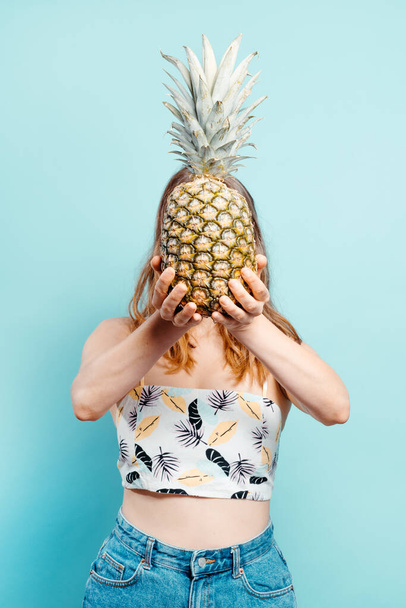 Cheerful young blonde holding a pineapple in front of her face with a summer top on a blue background. Summer concept. Vertical image with copy space - Photo, Image