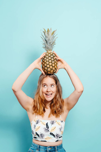 Fröhliche junge Blondine mit einer Ananas auf dem Kopf und einem Sommertop auf blauem Grund. Sommerkonzept. Vertikales Bild mit Kopierraum - Foto, Bild