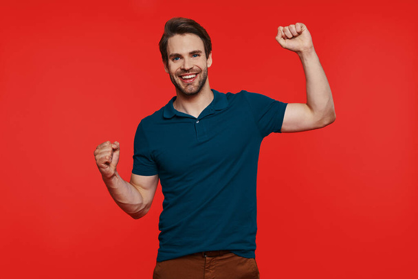 Happy young man in casual wear looking at camera and gesturing while standing against red background - Foto, Bild