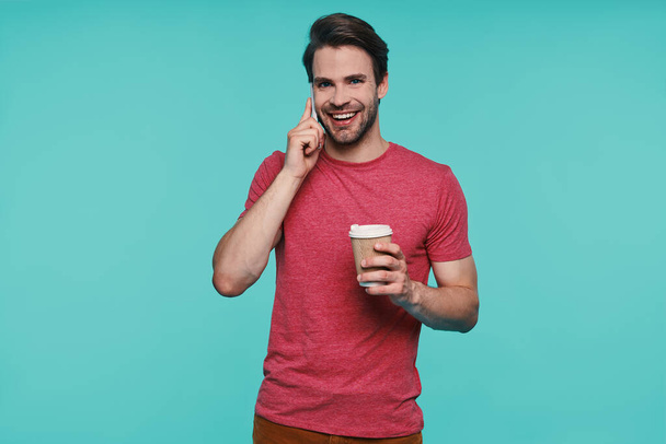 Handsome young smiling man in casual clothing holding cup and talking on smart phone - Foto, imagen